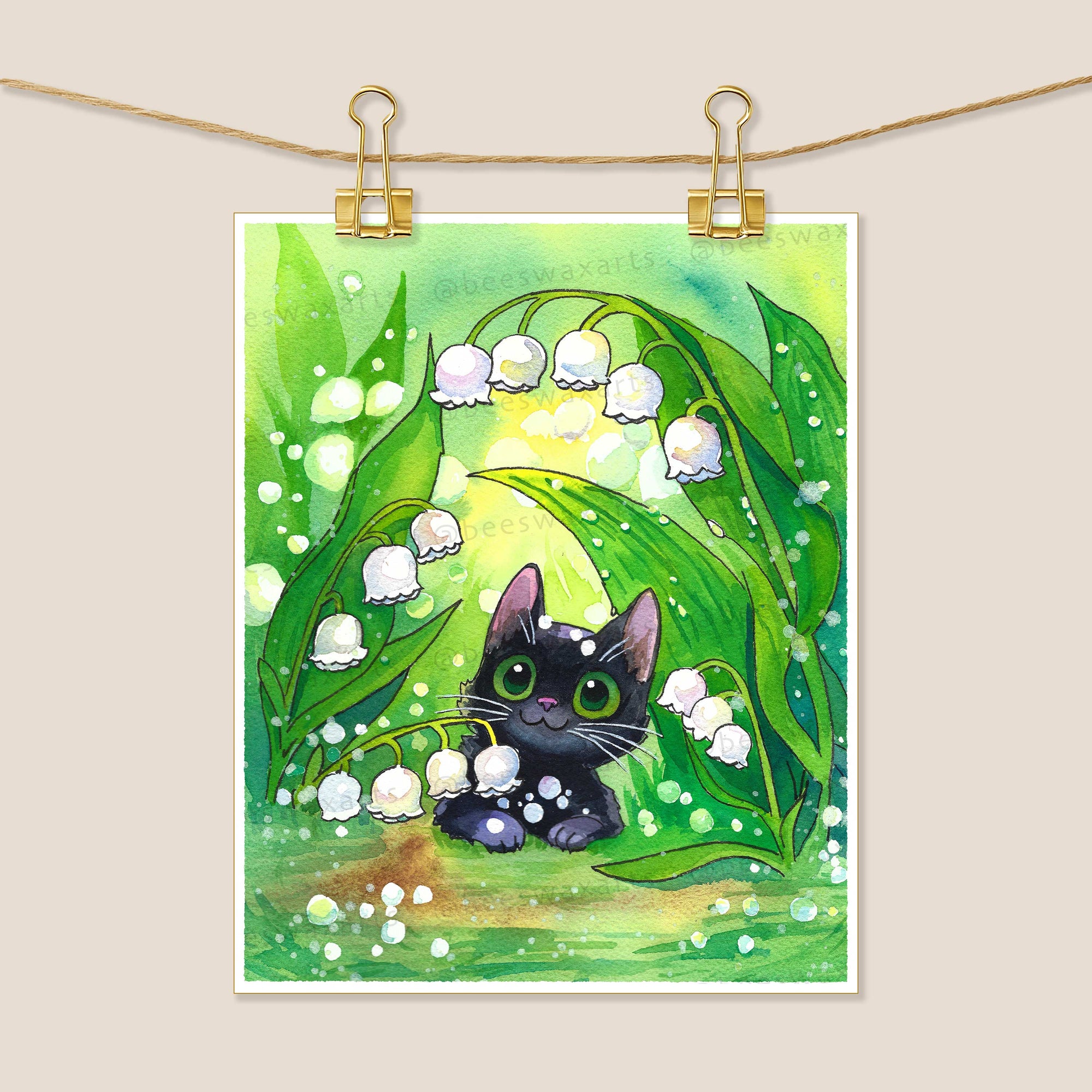 The Black Cat with Lilies of the Valley print by Beeswax Arts Watercolors features a green-eyed black cat among lily of the valley blooms, displayed on Arches Aquarelle paper and clipped with gold clips against a beige background.