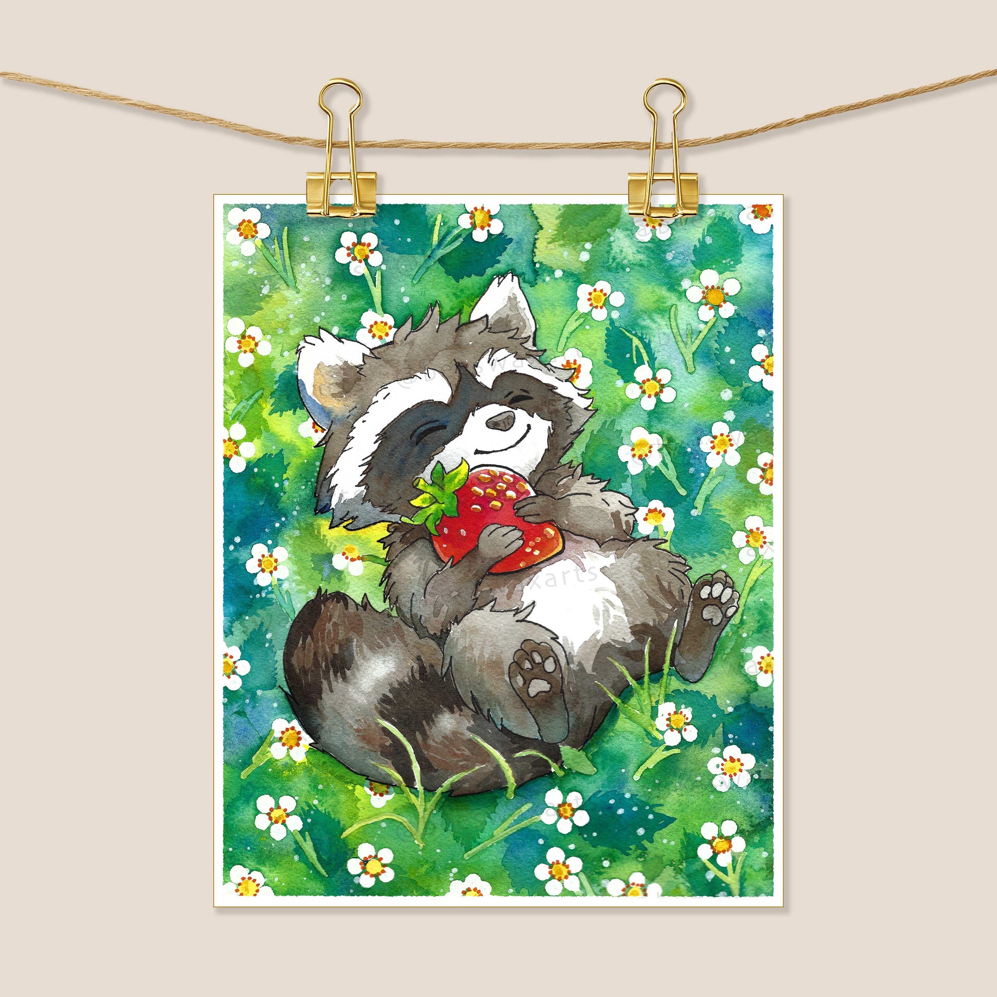 Raccoon with Strawberry by Beeswax Arts Watercolors features a raccoon lying in daisies, holding a red strawberry. This charming watercolor print is shown clipped to a string, with green grass and flowers in the background.