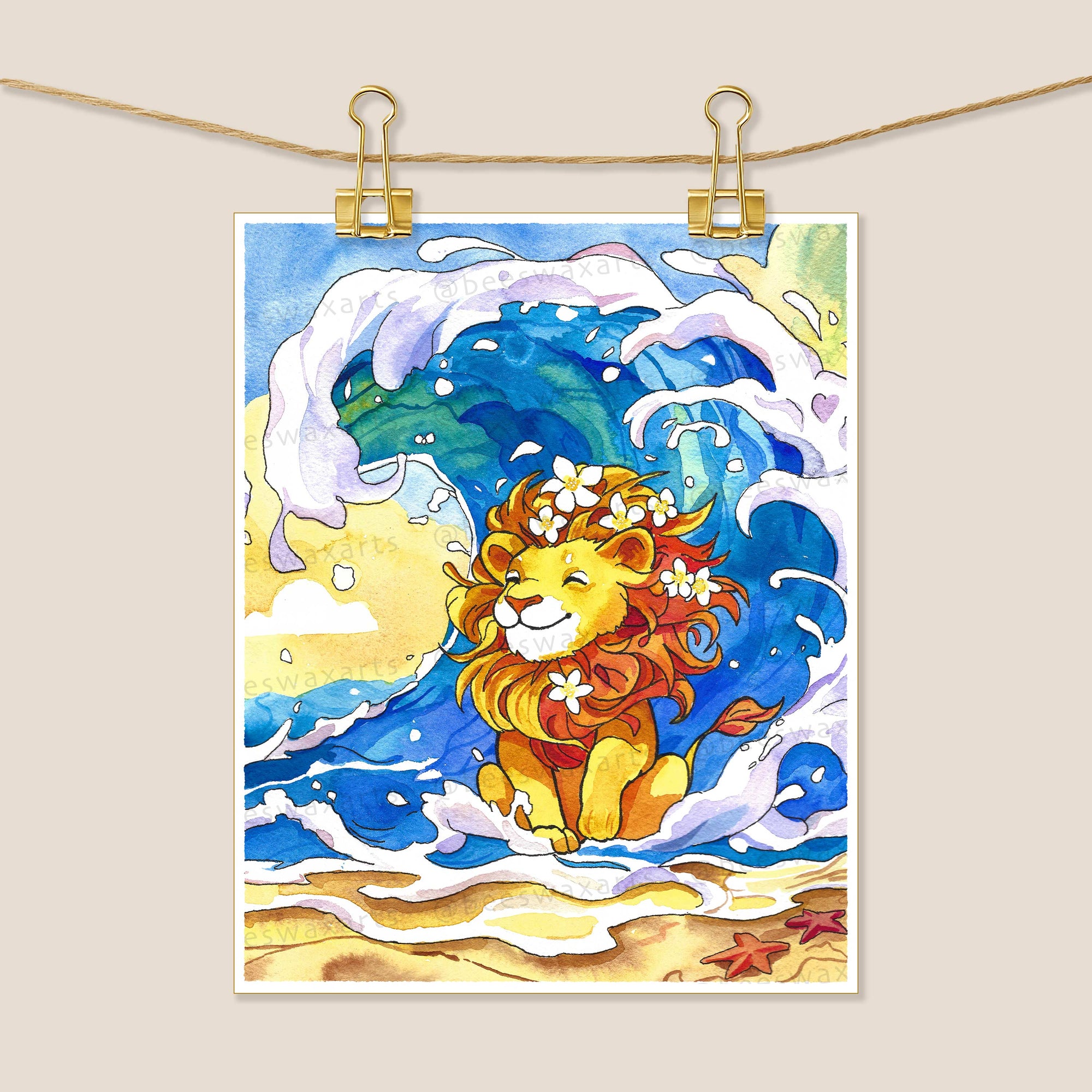 Beeswax Arts Watercolors Sea Lion art print features a cheerful lion with flowers in its mane on a sandy beach with a curling wave and two red starfish, displayed hanging by clips on a string.