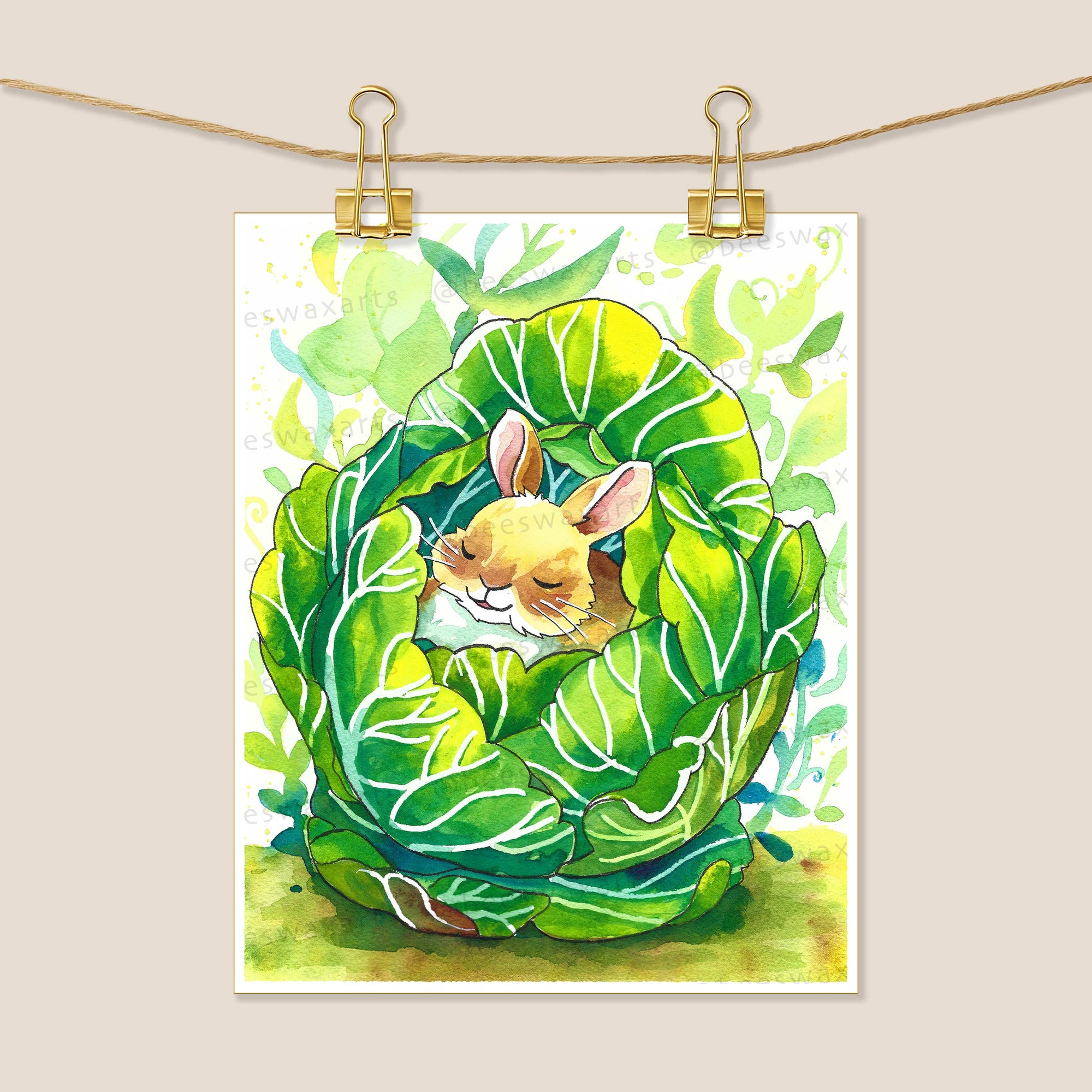 The Baby Bunny in a Cabbage 8x10 art print by Beeswax Arts Watercolors features a small brown bunny sleeping inside a leafy green cabbage. Shown with two gold binder clips, its perfect for framing and adds charm to any space.