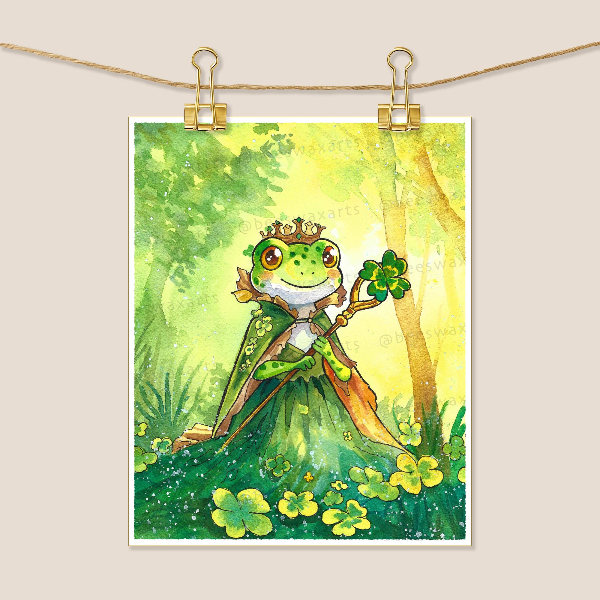 The Frog Princess Bringing Good Luck art print by Beeswax Arts Watercolors features a cheerful crowned frog with a four-leaf clover in a sunny, clover-filled forest, beautifully rendered in watercolor on premium archival paper.