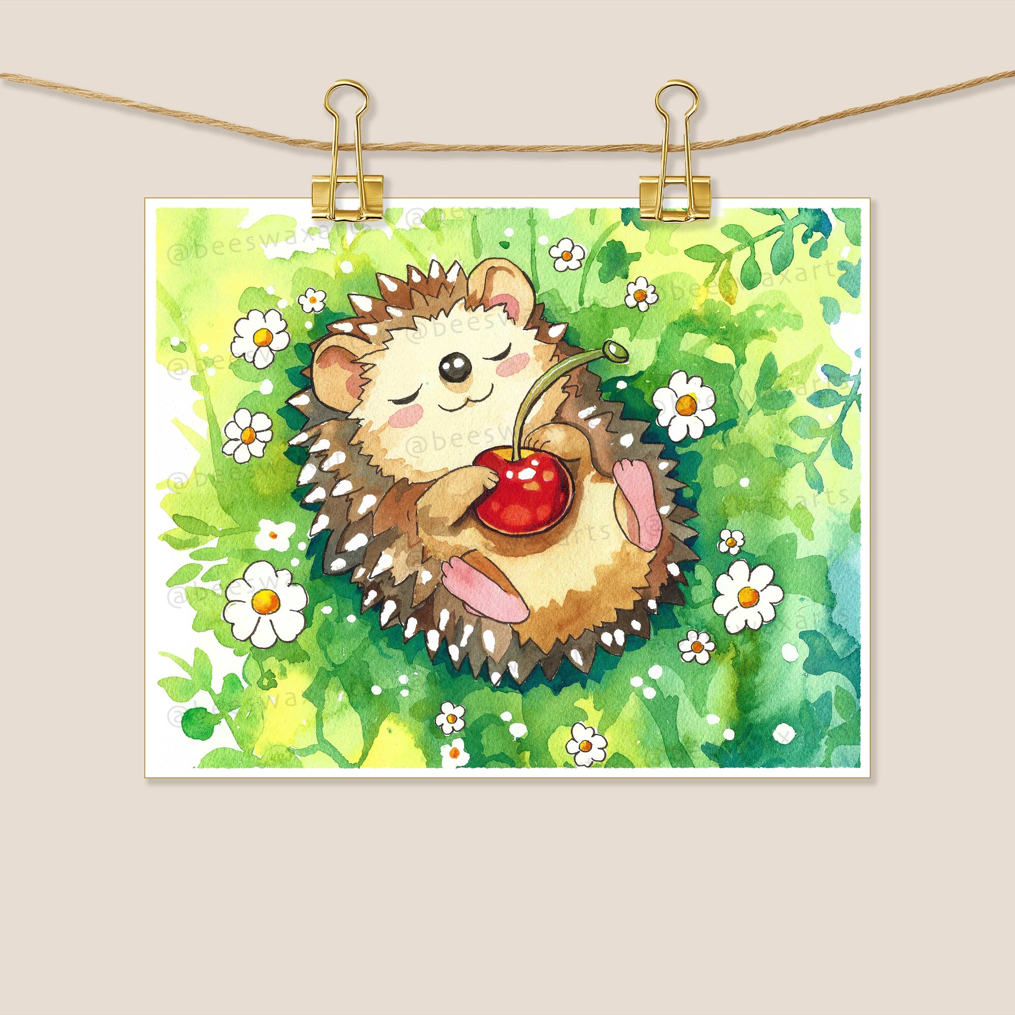 The Hedgehog cherry by Beeswax Arts Watercolors features a hedgehog lying on its back in green grass with white flowers, holding a red cherry. The watercolor artwork is clipped to a string with gold binder clips.