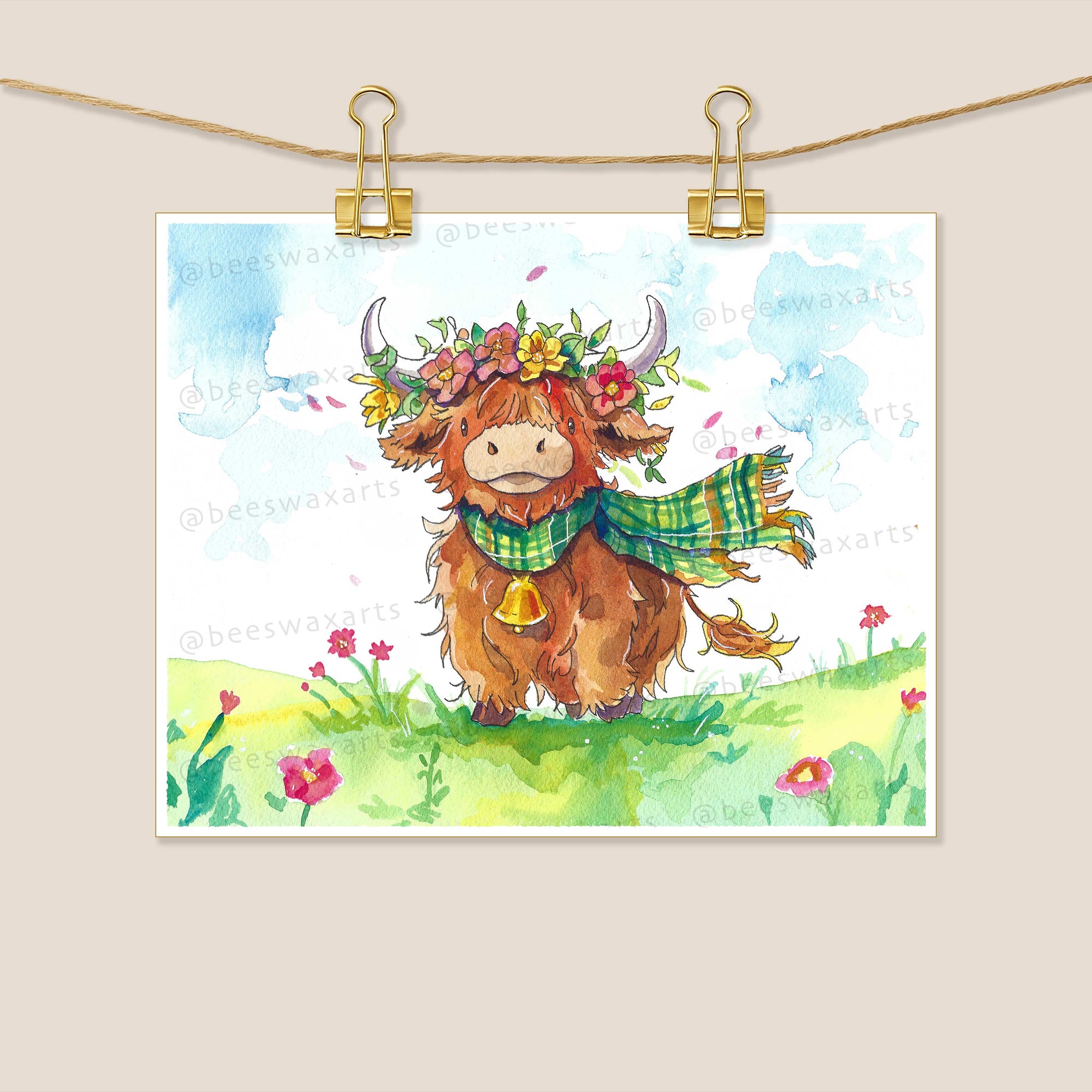 The Highland Cow by Beeswax Arts Watercolors features a smiling brown cow and fluffy alpaca in green plaid scarves and flower crowns, standing on grassy pink flowers under a blue sky, clipped to a string with gold clips.