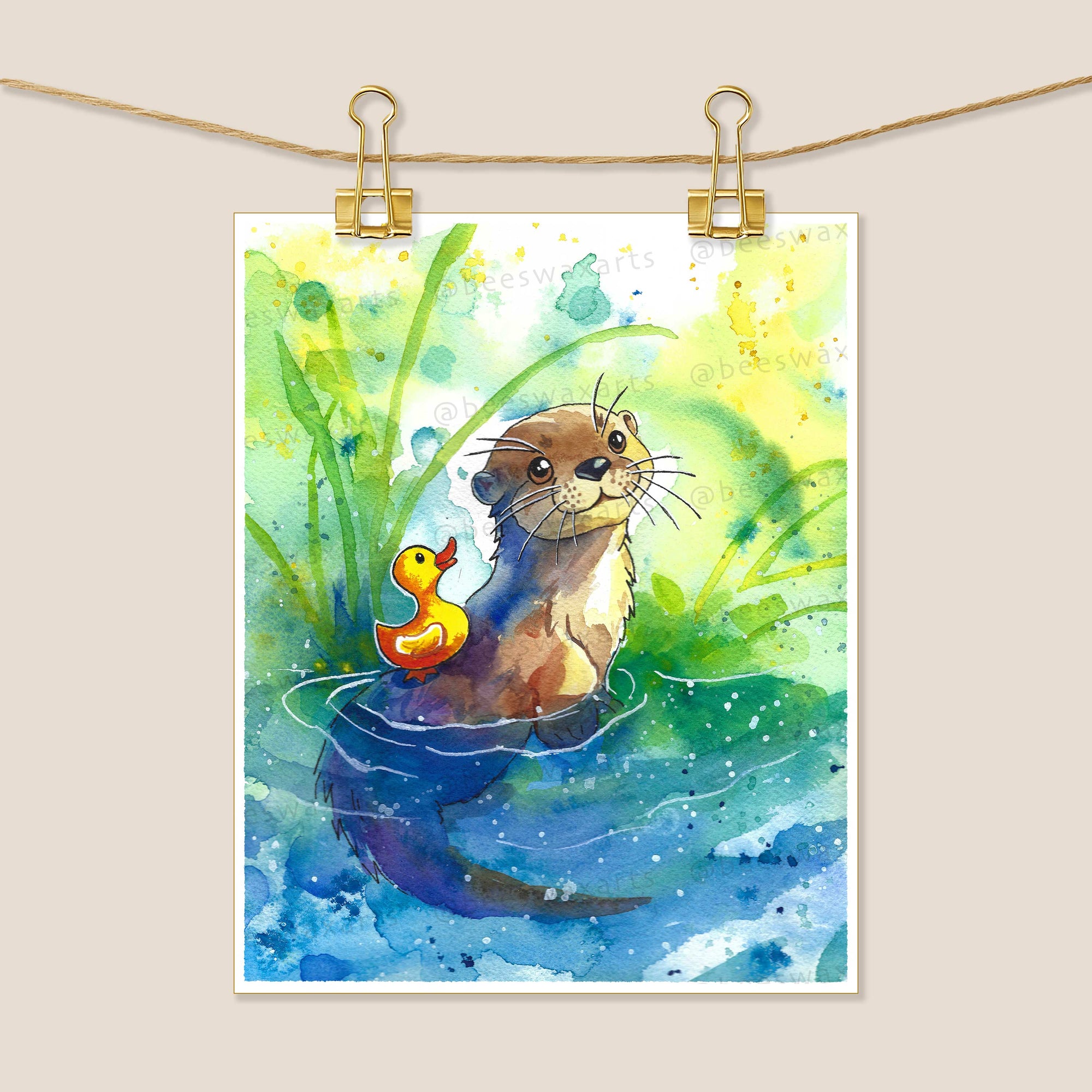 The River Otter and Rubber Duckie 8x10 art print by Beeswax Arts Watercolors features a watercolor otter with a yellow rubber duck, surrounded by green plants and a blue-yellow background, displayed clipped to a string with gold clips.